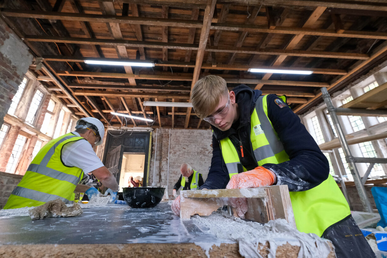 A person in a high-vis jacket, googles and gloves, creatging a plaster mould.