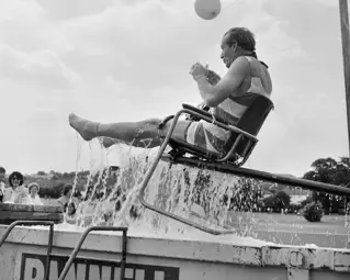 A man on a chair being lifted out of a skip filled with water.